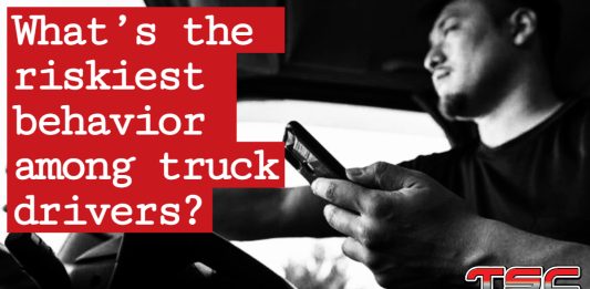 A professional truck driver operating a semi-truck while holding a cell phone, illustrating the rise in risky driving behaviors among truckers.